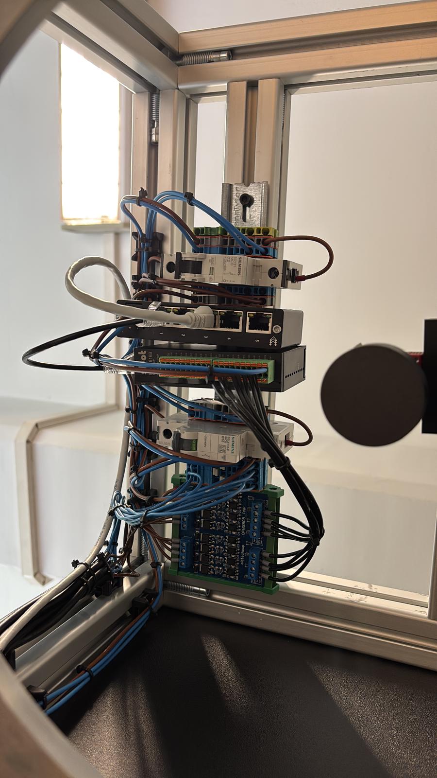 Control wiring on a DIN rail inside an aluminium-profile frame: circuit breakers, network switch, terminal blocks and a relay board